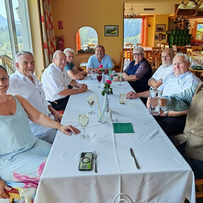 Eine Gruppe älterer Menschen sitzt an einem langen Tisch in einem Restaurant, lächelt und posiert für ein Foto. Der Tisch ist mit Weingläsern, Blumen und Besteck gedeckt. Im Hintergrund ist ein Blick auf Berge und einen Feuerlöscher zu sehen.