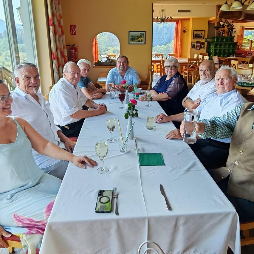 Eine Gruppe älterer Menschen sitzt an einem Tisch in einem Restaurant, lächelt und posiert für ein Foto. Der Tisch ist mit Weingläsern, Blumen und Tellern gedeckt. Im Hintergrund befinden sich weitere Tische und Stühle.