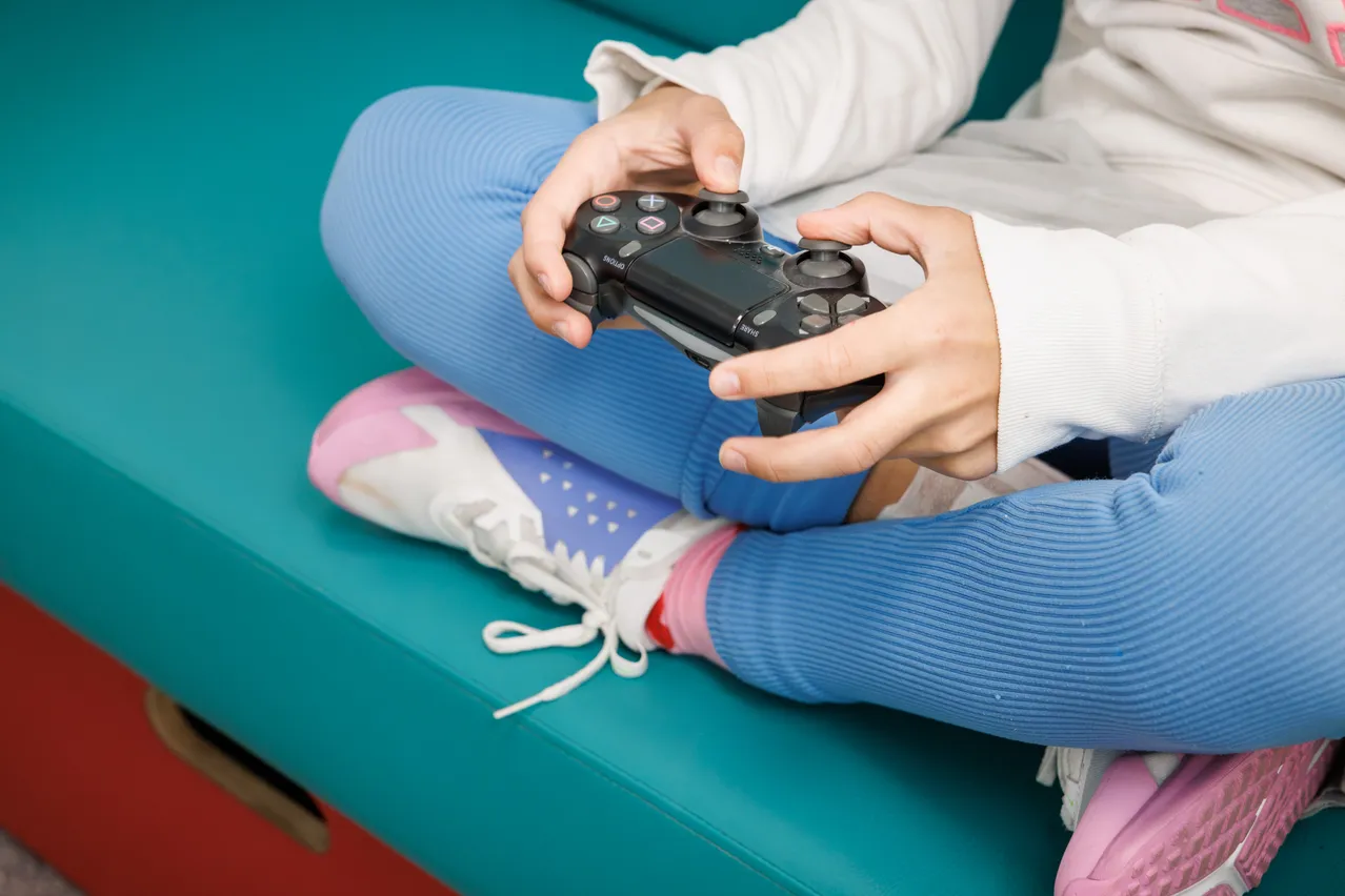 Eine Person sitzt auf einem blauen Sofa und hält einen schwarzen PlayStation-Controller in beiden Händen.