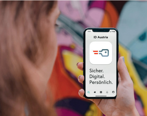 Eine Person hält ein Smartphone mit der ID Austria App und zeigt die Benutzeroberfläche und den Slogan der App. Der Bildschirm der App zeigt 'Sicher. Digital. Persönlich.' vor einem farbenfrohen Graffitihintergrund.