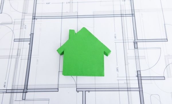 Ein grünes Papierhausmodell liegt auf einem detaillierten Architekturplan und zeigt seine Abmessungen und Anordnung.