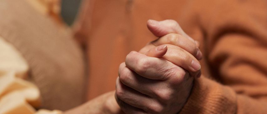 Ein älteres Paar, beide lächelnd, halten sich an den Händen, während sie zusammen tanzen. Die Frau trägt eine beige Weste, und der Mann trägt einen braunen Pullover und Hemd. Sie befinden sich wahrscheinlich in einer sozialen Umgebung.