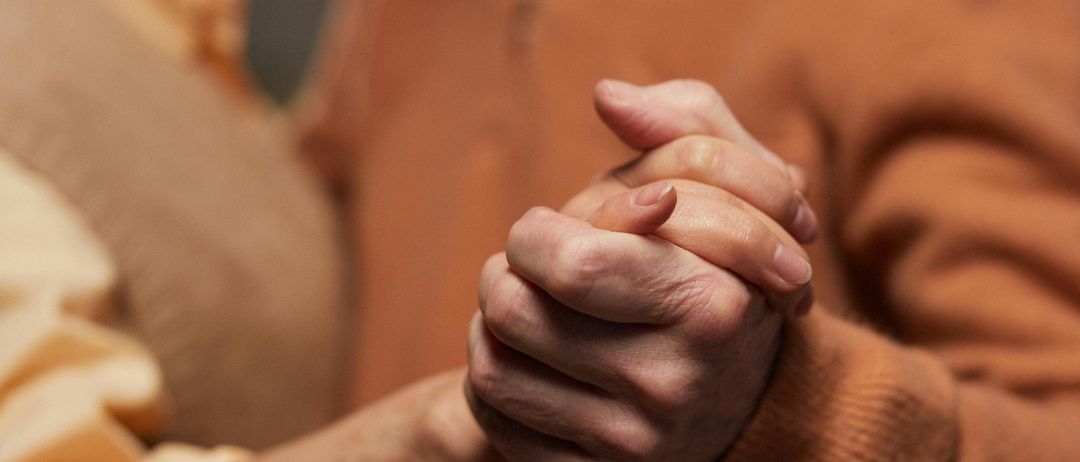 Ein älterer Mann und eine Frau halten sich an den Händen und lächeln, wobei der Mann einen braunen Pullover und die Frau ein beiges Oberteil trägt.