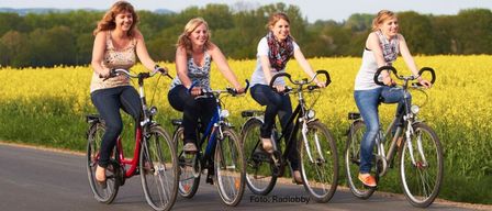 Fünf Frauen fahren an einem sonnigen Tag mit dem Fahrrad. Sie lächeln und scheinen ihre Fahrt zu genießen. Sie fahren durch ein Feld mit gelben Blumen.