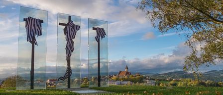 Drei Glaskreuze mit drapiertem schwarz-weiß gestreiftem Stoff hängen auf einer Rasenfläche mit einem bewölkten Himmel im Hintergrund.