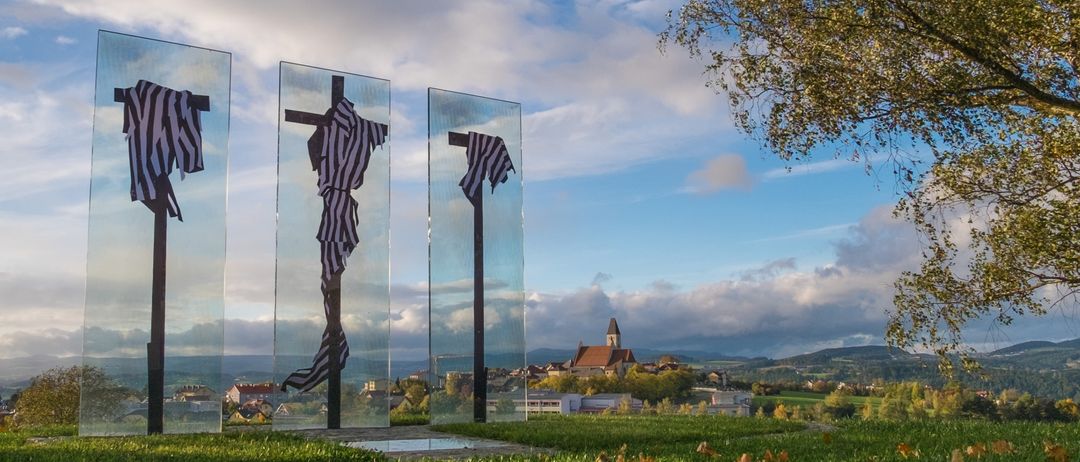 Drei Glaskreuze mit drapiertem schwarz-weiß gestreiftem Stoff hängen auf einer Rasenfläche mit einem bewölkten Himmel im Hintergrund.
