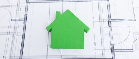 Ein grünes Hausmodell liegt auf einem blauen architektonischen Plan. Das Hausmodell ist einfach, mit einem dreieckigen Dach. Der Plan zeigt verschiedene Linien und Maße.