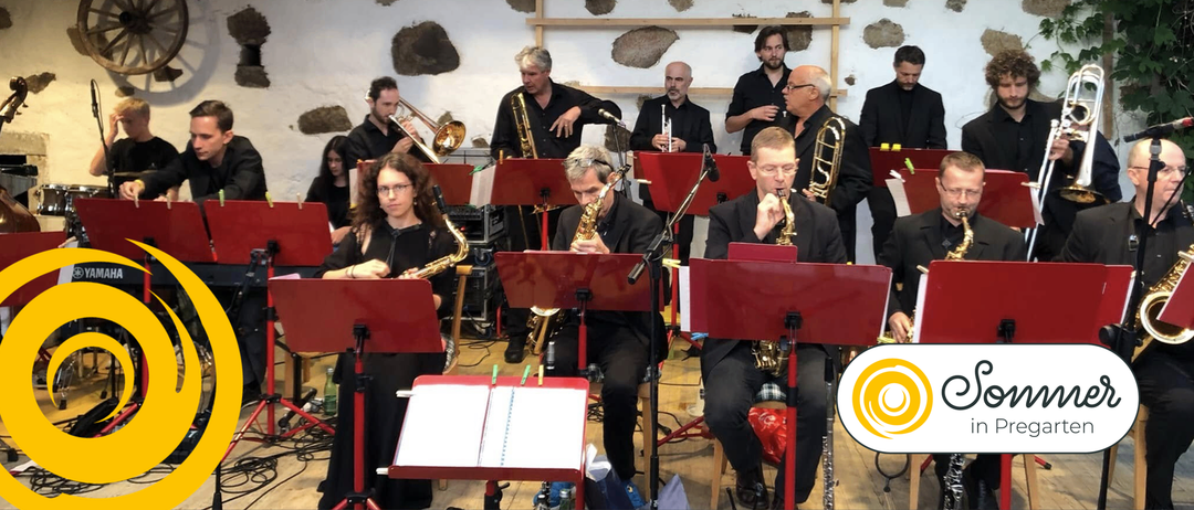 Eine Gruppe von Musikern spielt Instrumente, darunter Posaunen, Trompeten und Saxophone. Sie sitzen auf Stühlen, einige stehen. Sie haben Notenständer vor sich.