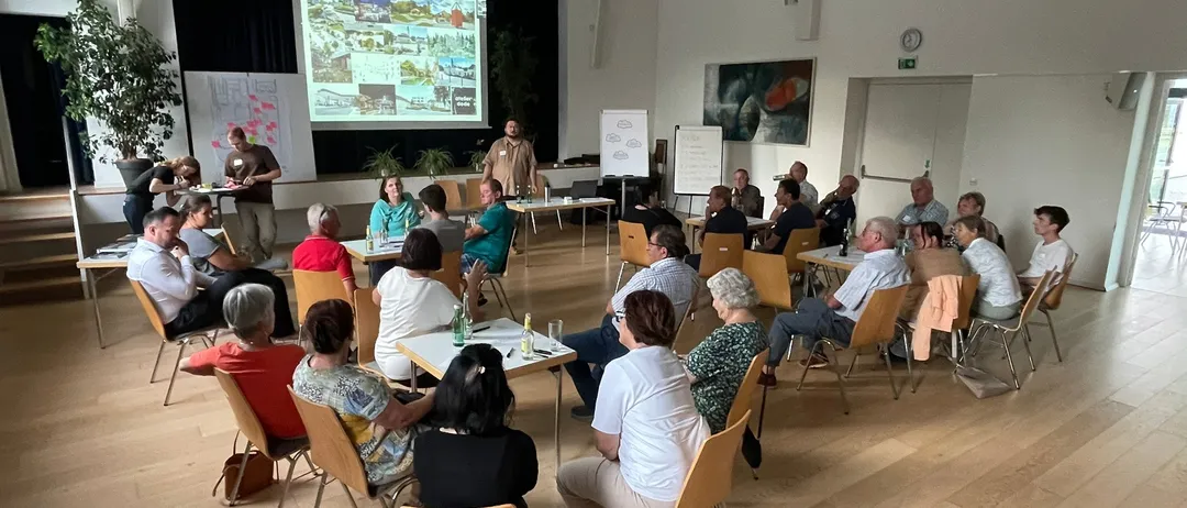 Eine Gruppe von Menschen sitzt in Stühlen in einem Raum und hört sich eine Präsentation auf einer Projektionsleinwand an.