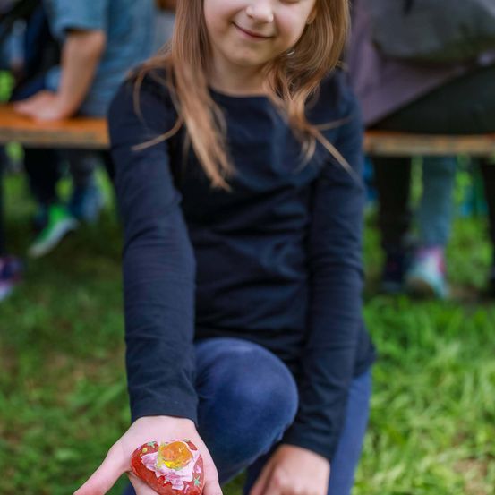 Ein Mädchen hält einen herzförmigen Gegenstand in der Hand und lächelt, während sie ihn ansieht. Ein Mensch sitzt hinter ihr auf einer Bank.
