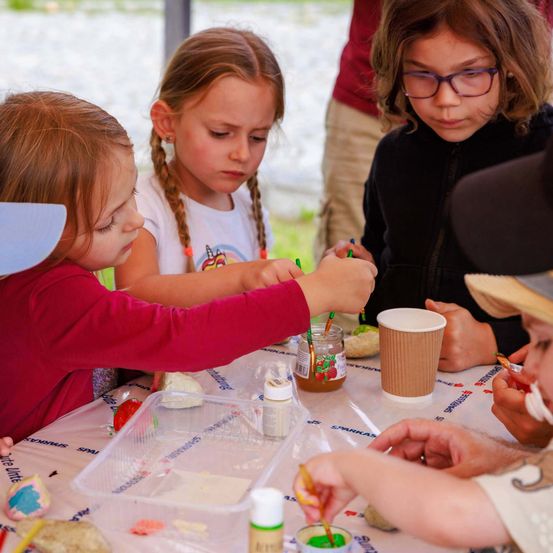 Kinder sind bei einer Kunstaktivität am Tisch beschäftigt, sie malen oder dekorieren Objekte. Ein Becher und ein Glas stehen auf dem Tisch.