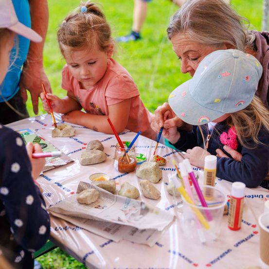 Eine Gruppe von Kindern und Erwachsenen malt Steine an einem Tisch im Freien. Der Tisch hat Farbe, Pinsel und Becher. Ein Kind hat eine Regenbogenmütze.