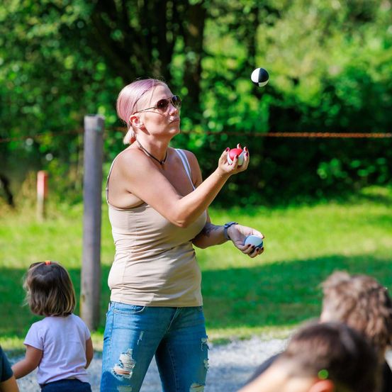 Eine Frau mit rosa Haaren jongliert mit zwei Bällen, einem roten und einem weißen, in einem grasbewachsenen Außenbereich. Sie trägt eine Sonnenbrille und eine Halskette. Ein kleines Mädchen steht hinter ihr, und mehrere Menschen beobachten sie.