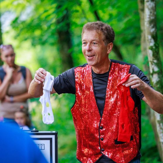 Ein Mann in einem roten Paillettenweste und schwarzem T-Shirt führt draußen in einem Wald einen Zaubertrick mit einer weißen Socke vor.