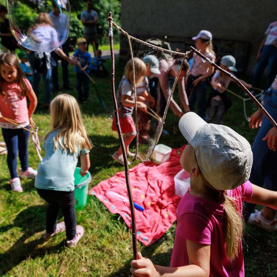 Mehrere junge Mädchen spielen mit großen Seifenblasen auf einem Rasen. Ein Mädchen hält einen Stock mit einem Netz. Andere Kinder sind in der Nähe und halten ähnliche Stöcke. Einige pusten Seifenblasen. Ein Tisch mit Wasser steht in der Nähe.