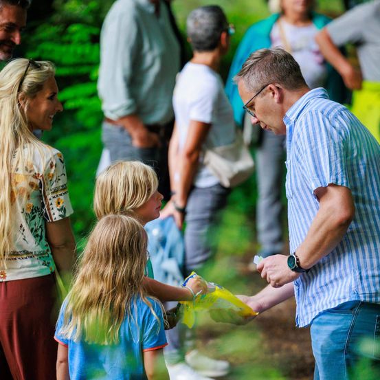 Eine Gruppe von Menschen in einem Park, wobei ein Mann einem jungen Mädchen eine gelbe Tasche gibt.
