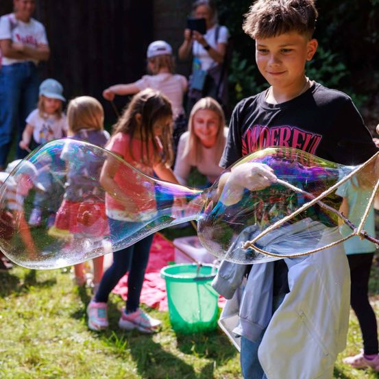 Ein Junge bläst einen riesigen Seifenblasen im Freien, umgeben von anderen Kindern, während Erwachsene zusehen und einige Kameras halten.
