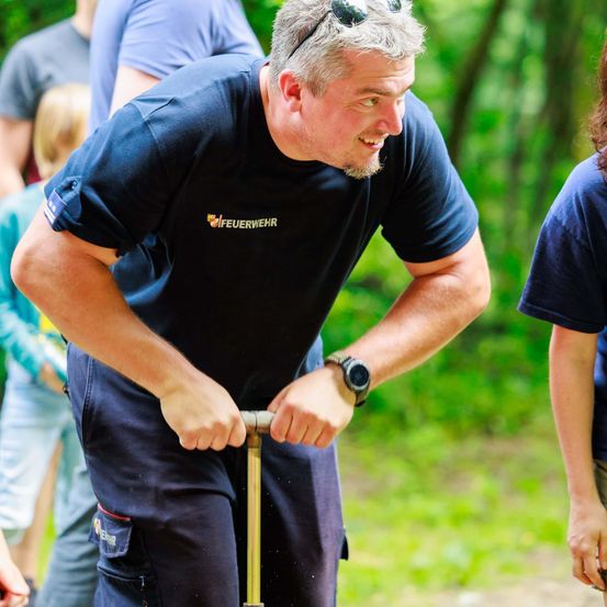 Ein Feuerwehrmann in blauer Uniform fährt einen Roller, lächelt und schaut nach vorne. Hinter ihm stehen ein Kind und eine weitere Person. Der Hintergrund ist ein verschwommener Wald.
