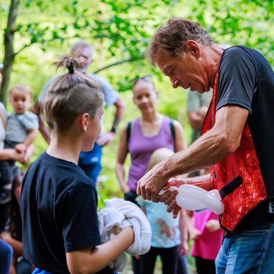 Ein Mann in einem roten Vest bläst einem Jungen mit einem Handtuch Luftballons auf, während andere in einem Wald zuschauen.