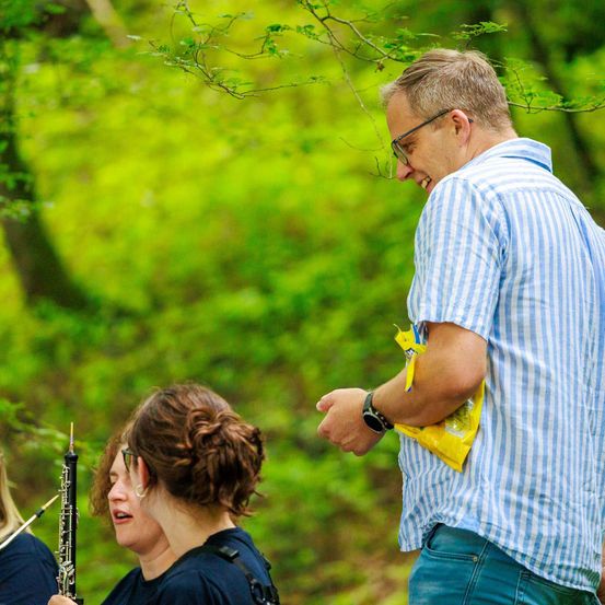 Ein Mann mit Brille lächelt, während er eine Frau beobachtet, die eine Klarinette spielt. Eine weitere Frau steht hinter ihr, alle befinden sich in einem Wald.
