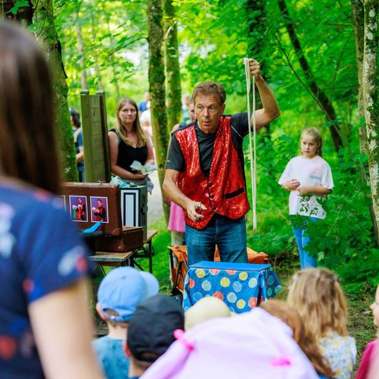Ein Zauberer führt in einem Wald einen Seiltrick vor, umgeben von Kindern und Zuschauern. Der Zauberer trägt ein rotes Paillettenweste und hält ein Seil. Ein Mädchen in einem rosa Shirt steht in der Nähe.