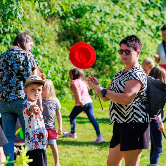 Eine Frau mit einem roten Ballon dirigiert einen jungen Jungen in einem grasigen Außenbereich, umgeben von anderen Kindern und Erwachsenen.