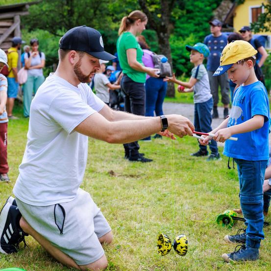 Ein Mann hockt und hält eine Schnur und beschäftigt einen Jungen in einem Spiel. Sie sind von anderen Zuschauern umgeben, mit Bäumen und einem Gebäude im Hintergrund.
