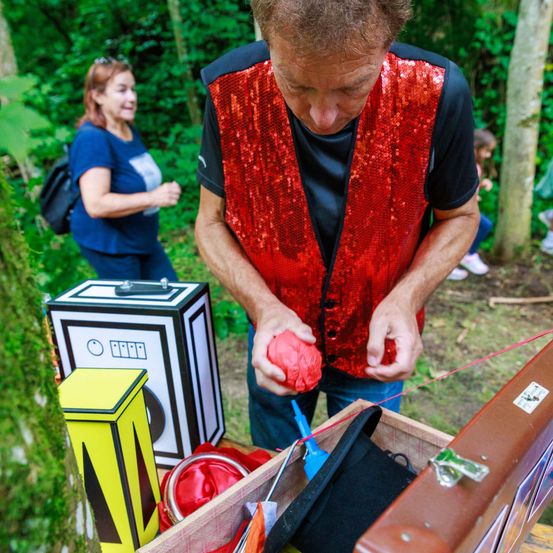 Ein Mann in einem roten Paillettenweste schaut sich einen roten Ball an, steht in einem Wald mit einem Koffer voller Requisiten vor sich. Eine Frau in Blau geht hinter ihm.