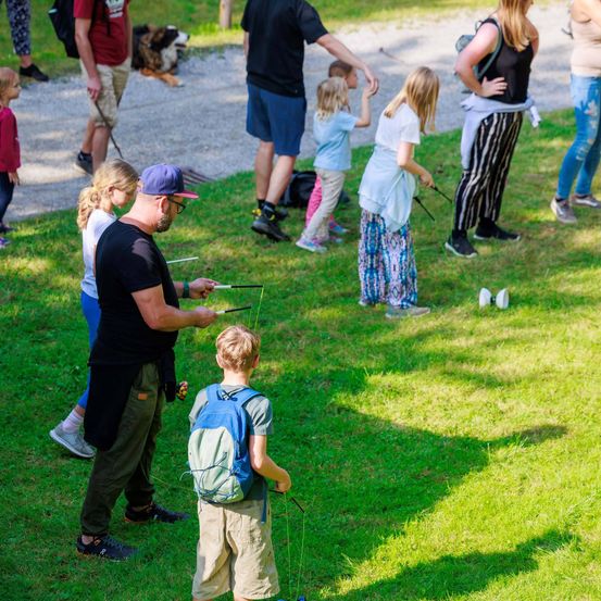 Eine Gruppe von Menschen versammelt sich auf einem Rasenfeld, einige fischen, während andere zuschauen. Kinder spielen im Hintergrund.
