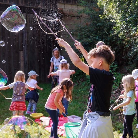 Kinder spielen draußen an einem sonnigen Tag. Ein Junge hält einen Stab mit einer Seifenblasenschlaufe und erzeugt große Seifenblasen. Andere Kinder beobachten und halten ihre eigenen Seifenblasenschlaufen. Eine Frau steht hinter ihnen, und ein Eimer und eine Schüssel liegen auf dem Boden.