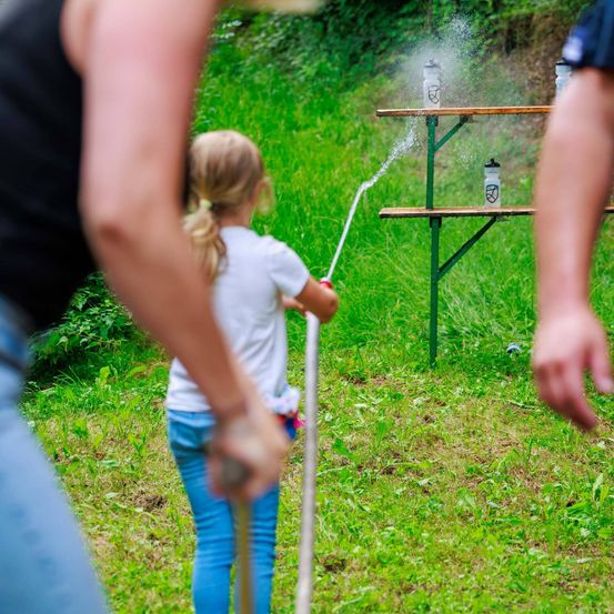Ein Mädchen mit Zöpfen sprüht Wasser aus einem Schlauch auf einen Tisch mit Flaschen. Hinter ihr hält eine Person den Schlauch. Im Hintergrund steht eine weitere Person in der Nähe des Tisches.