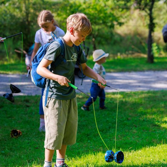 Ein Junge spielt im Park mit einem Spielzeug. Er hält eine grüne Schnur mit einem Spielzeug am Ende. Hinter ihm sind ein Mädchen und ein Baby, das einen Hut trägt.