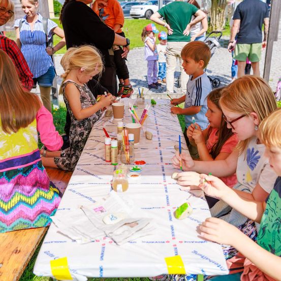 Kinder malen an einem Tisch im Freien, mit Erwachsenen und anderen Kindern in der Nähe. Der Tisch hat Becher, Farbe und andere Malmaterialien.