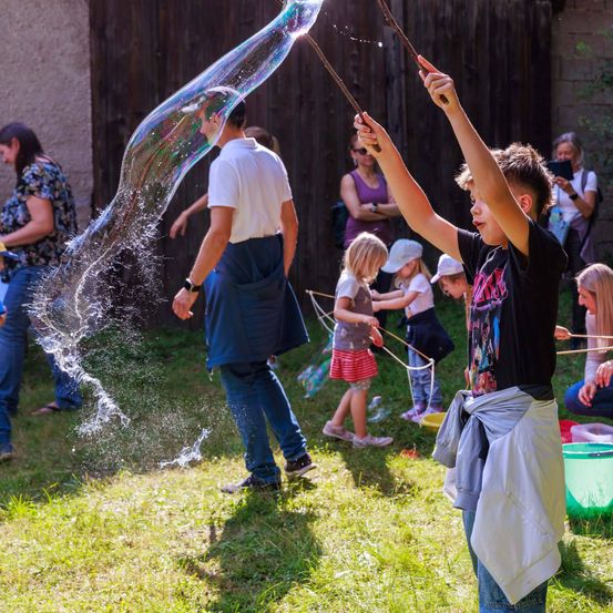 Ein Junge bläst mit einem Stock eine riesige Seifenblase, während andere Kinder mit Blasen spielen. Ein Mann und eine Frau stehen in der Nähe, und mehrere andere Personen sind im Hintergrund.