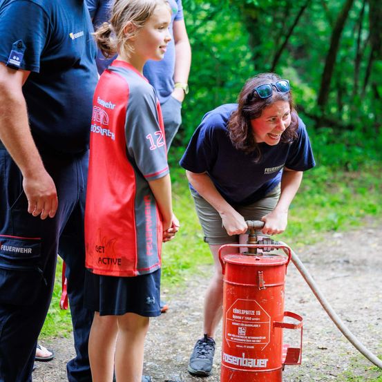 Eine Frau benutzt einen Feuerlöscher, während ein junges Mädchen und zwei Männer in einem Außenbereich mit grünen Bäumen zusehen.