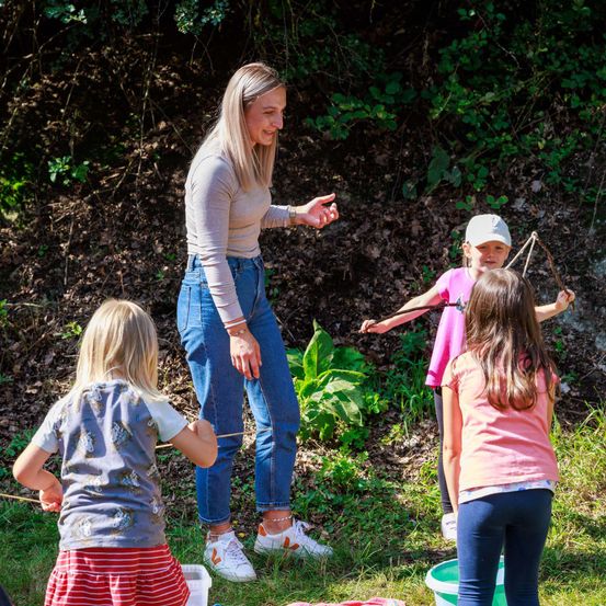 Eine Frau steht mit drei jungen Mädchen in einem Grasbereich. Sie halten Stöcke und scheinen zu spielen.