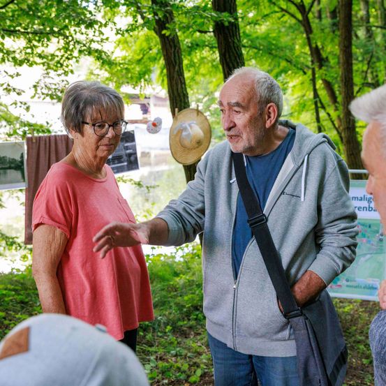 Ein älterer Mann in einer grauen Kapuzenjacke spricht mit einer älteren Frau in einem rosa T-Shirt, beide stehen in einem Wald. Hinter ihnen ist eine Karte an einem Baum befestigt.