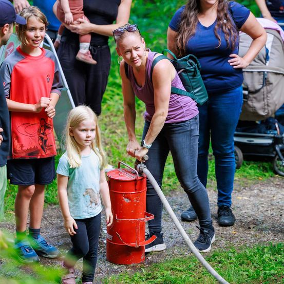 Eine Frau pumpt Wasser aus einem Fass, während ein Mädchen zuschaut, umgeben von anderen Menschen, einige mit Kindern, in einem Park.