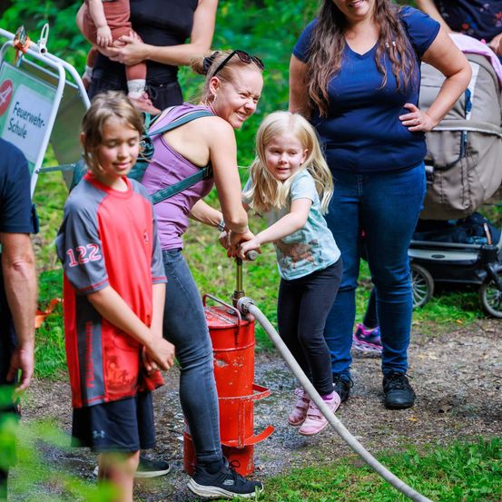Eine Frau und ein junges Mädchen pumpen Wasser aus einer roten Pumpe, während andere in einem Park zuschauen.