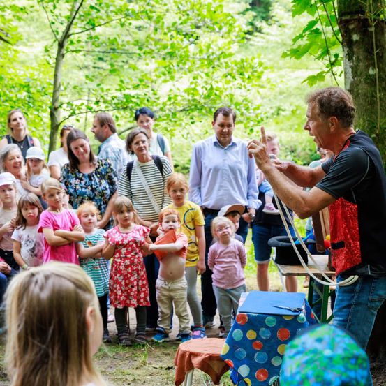 Eine Gruppe von Menschen, einschließlich Kindern, versammelt sich in einer Waldlichtung. Ein erwachsener Mann, möglicherweise ein Zauberer, führt einen Trick mit einem Seil vor ihnen durch.