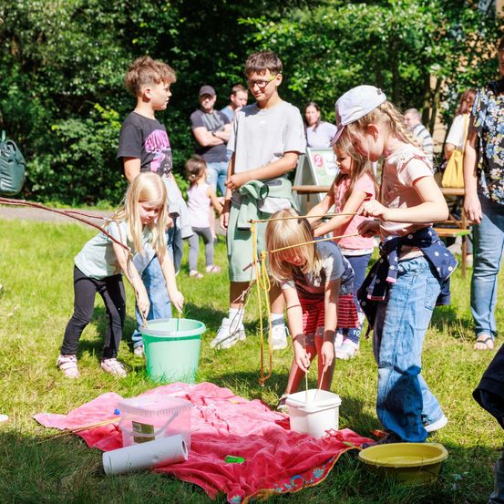 Kinder in einem Park sind in einer verspielten Aktivität engagiert und verwenden Stöcke, um Wasser aus Eimern zu übertragen. Sie stehen auf einer Decke mit Eimern und einer gelben Schüssel. In der Nähe beobachten andere und stehen auf Gras.