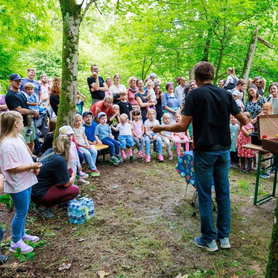 Ein Mann unterhält eine Gruppe von Kindern und Erwachsenen im Wald. Einige sitzen, andere stehen. Flaschen mit Wasser stehen auf dem Boden.