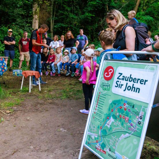 Eine Gruppe von Menschen ist in einem Waldgebiet versammelt, wobei eine Person vor ihnen auftritt. Ein Schild mit einer Karte ist auf dem Boden platziert.