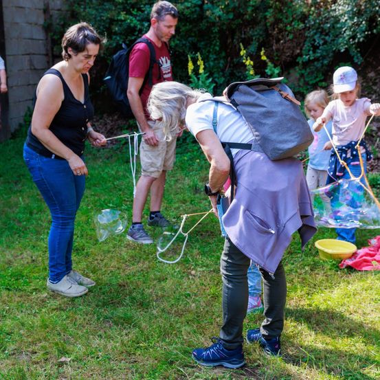 Eine Gruppe von Erwachsenen und Kindern befindet sich im Garten. Sie halten Schnüre mit Seifenblasen. Es gibt Rucksäcke und eine gelbe Schüssel auf dem Rasen.
