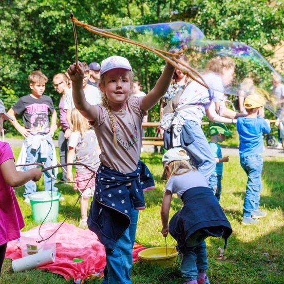 Eine Gruppe von Kindern spielt draußen an einem sonnigen Tag. Ein kleines Mädchen hält eine Seifenblasen-Wand und pustet eine Blase. Ein anderes Kind beugt sich vor. In der Nähe spielen Kinder mit einem Stock und einem Eimer. Bäume und Erwachsene sind im Hintergrund.