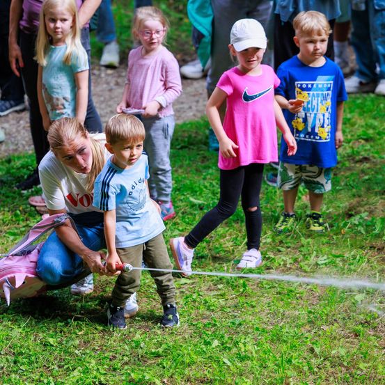 Eine Frau hilft einem kleinen Jungen, einen Schlauch auf einem Rasen zu benutzen, während andere Kinder und Erwachsene im Hintergrund zusehen.