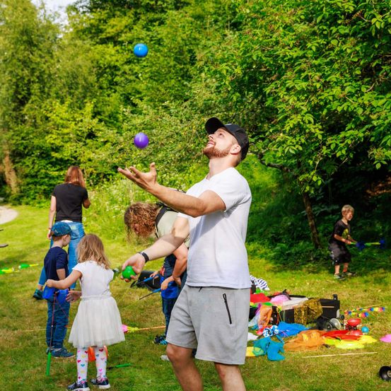 Ein Mann jongliert mit bunten Bällen in einem Park, umgeben von Kindern und Erwachsenen. Die Umgebung ist draußen mit Bäumen und einem Rasenfeld.