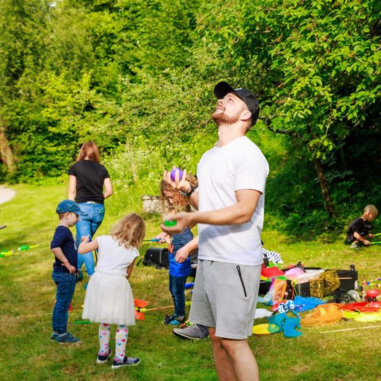 Eine Gruppe von Menschen im Freien, ein Mann jongliert mit bunten Bällen, Kinder spielen mit Spielzeug und eine Frau beobachtet. Bäume und Gras umgeben das Gebiet.