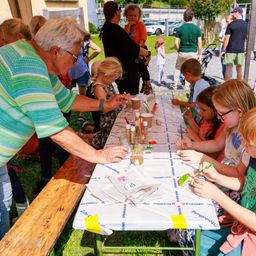 Eine Gruppe von Kindern malt auf einem Tisch in einem Park. Eine ältere Frau steht am Tisch und hilft den Kindern. Hinter dem Tisch stehen weitere Personen.