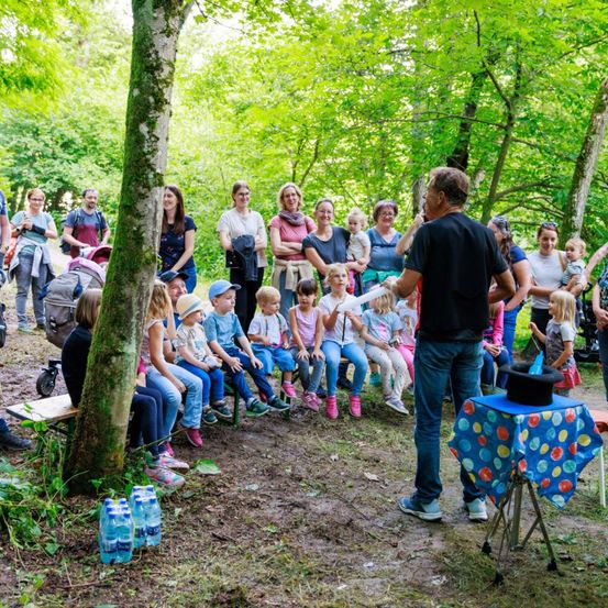 Ein Mann führt in einem Wald einen Zaubertrick für eine Gruppe von Kindern und Erwachsenen vor.
